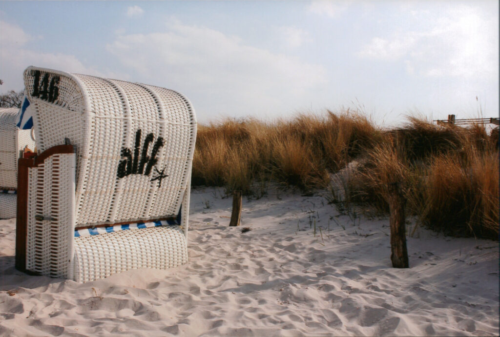 Ein einzelner, weiß geflochtener Strandkorb mit blauen Streifen steht prominent im weißen Sand eines Strandes, vermutlich an der Ostsee. Auf der Rückseite des Korbes ist in schwarzen Lettern das Wort 'Scharbeutz' mit einer stilisierten Krone geschrieben. Im Hintergrund erstrecken sich Dünen mit hohem, trockenem, braunem Dünengras unter einem hellen Himmel.