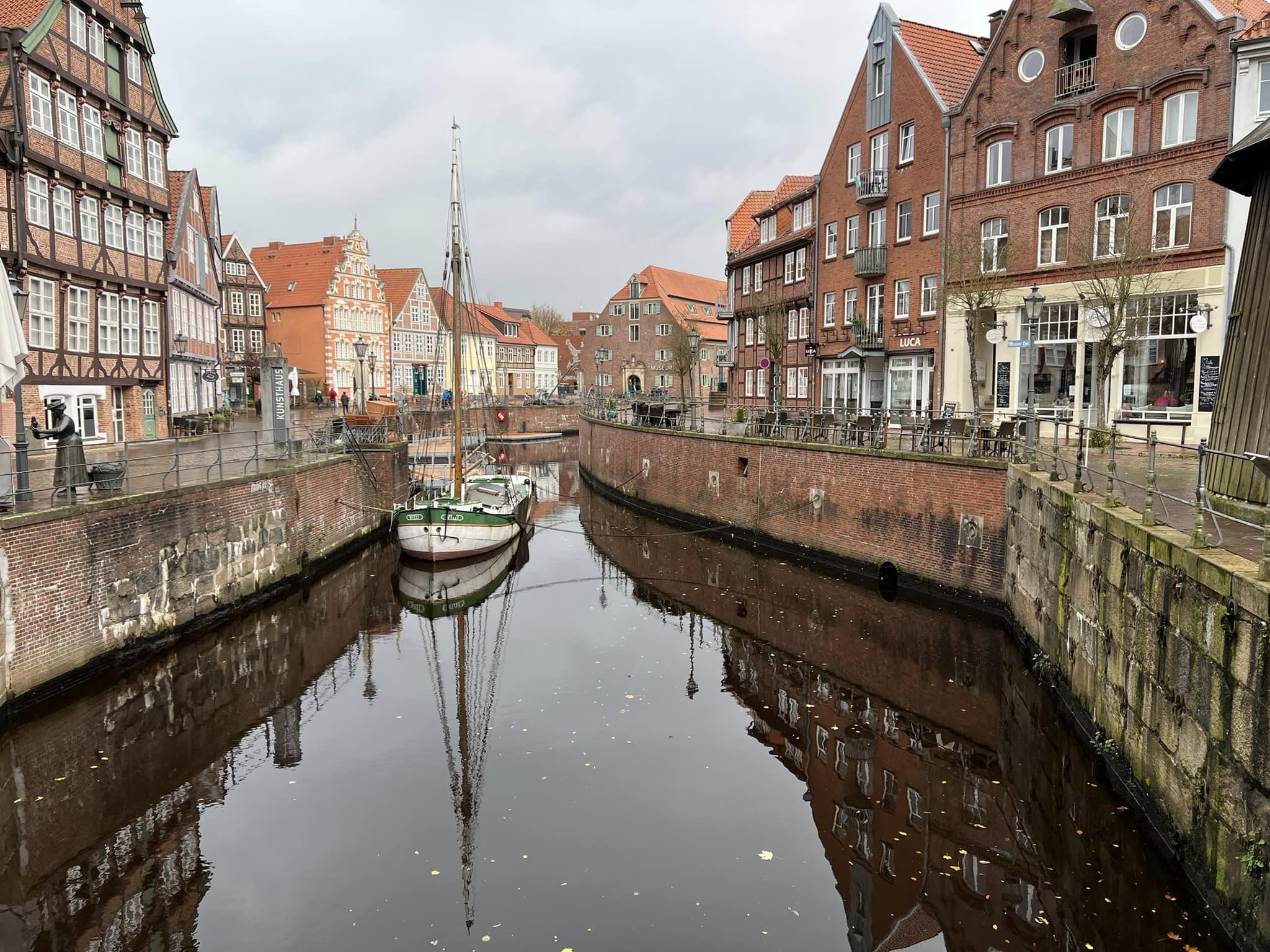 Eine Ansicht des Alten Hansehafens in Stade. Der schmale Kanal wird von hohen, roten Backsteinmauern und historischen Bürgerhäusern mit Fachwerkelementen und roten Ziegeldächern gesäumt. Ein traditionelles, kleines Segelschiff mit Mast liegt mittig im dunklen Wasser, dessen Spiegelung deutlich zu sehen ist. Der Himmel ist bedeckt, und die Ufer sind mit Tischen und Stühlen von Cafés ausgestattet.
