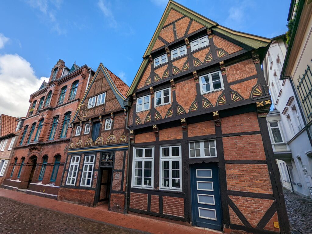 Eine Nahansicht von zwei gut erhaltenen, historischen Fachwerkhäusern in Stade, die mit dunkelbraunen Holzbalken und leuchtend roten Backsteinen verziert sind. Die Häuser haben spitze Giebel und weiße Fensterrahmen. Das rechte Haus zeigt dekorative Halbkreismuster im Fachwerk. Links stehen weitere ältere Backsteingebäude, und der Vordergrund zeigt eine nasse, gepflasterte Straße.