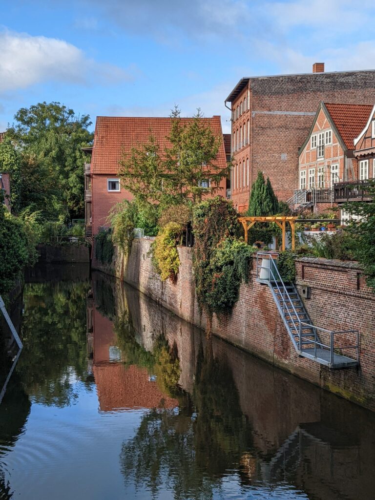 Eine ruhige Aufnahme eines schmalen Kanals, der von hohen Backsteinmauern und historischen, norddeutschen Fachwerkhäusern mit roten Ziegeldächern gesäumt wird. Die Gebäude und die Ufermauern spiegeln sich deutlich in der ruhigen Wasseroberfläche. Eine Metalltreppe führt von der Mauerkrone hinunter zu einer kleinen Plattform am Wasser. Das Ufer ist dicht mit Grünpflanzen und Bäumen bewachsen.