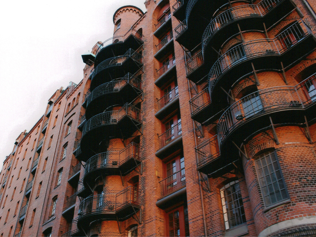 Untersicht eines hohen, historischen Speichergebäudes in Hamburg aus rotem Backstein. Die Fassade wird dominiert von einem zentralen, runden Turm mit mehreren halbrunden, schwarzen Eisenbalkonen, die sich übereinander stapeln. Die symmetrischen Fenster und die gotischen Giebel des Speichers sind ebenfalls gut sichtbar.