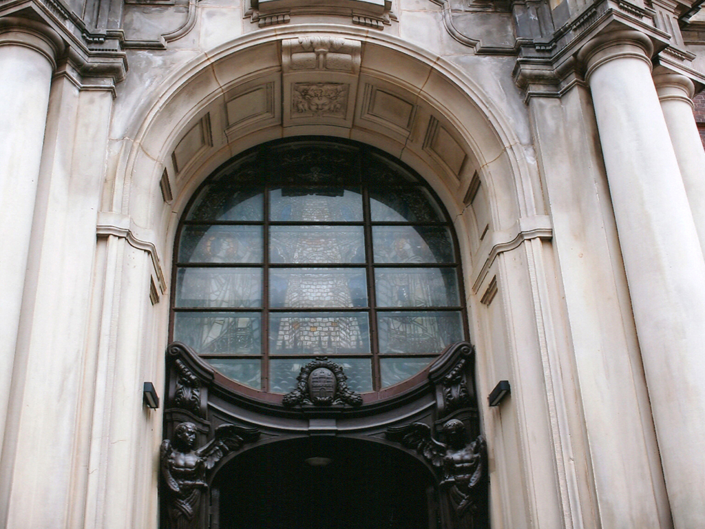 Nahaufnahme eines historischen Kirchenportals oder Gebäudeeingangs mit einer großen, halbkreisförmigen Buntglasfenster-Front. Das Portal ist von hellen, steinernen Säulen flankiert. Unter dem Fenster befindet sich eine reich verzierte dunkle Tür mit Reliefs von Engeln oder mythologischen Figuren. Das Fenster selbst ist in Metallrahmen unterteilt und zeigt möglicherweise eine christliche Figur.